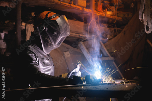 A male welder in a leather jacket carries out welding work in a home shed against the background of a brick wall. Handicraft production of metal structures and products