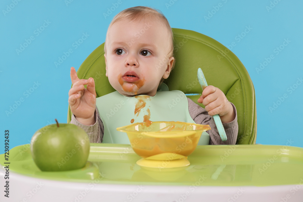 Cute little baby eating healthy food in high chair on light blue background Cute little baby eating healthy food in high chair on light blue background