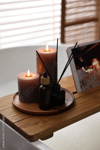 Wooden tray with burning candles, aroma diffuser and essential oil on bathtub in bathroom