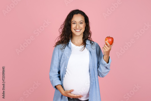 Young pregnant woman eating apple on pink background