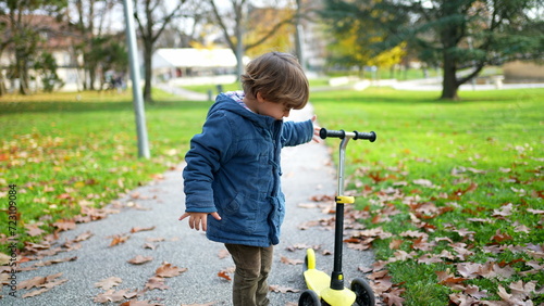 One little boy standing at park during autumn day wearing blue jacket checking his 3 wheeled scooter
