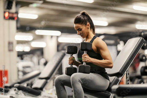 A strong woman is sitting in a gym and exercising with dumbbells.