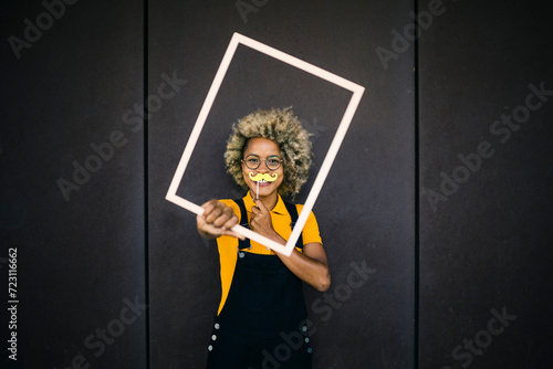 Woman holding fake mustache and frame in front of black wall photo