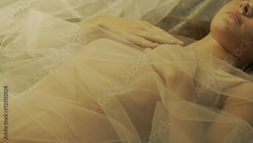 Hands of woman with nude manicure, folded on woman's chest, close up. Woman lying on her back covered with white tulle, illuminated by warm rays of light. Slow motion. photo