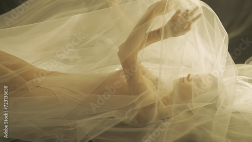 Side view of a young woman on a bed, under a transparent air cloth, close up. A woman illuminated by warm light raised her arms, forming a tulle like canopy.