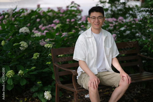 smile Chinese young man sit on park bench with flowers behind in summer