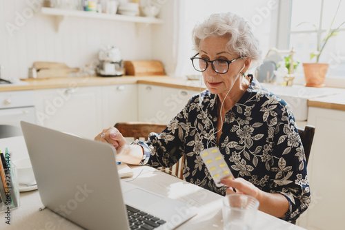 Telemed concept. Indoor portrait of old woman in earphones holding blister package of pills in hands and showing it ti laptop camera consulting with her doctor online, asking for prescriptions photo