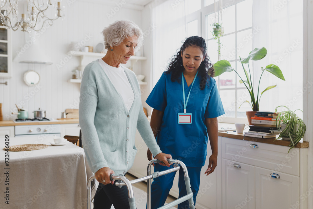 Side view of black african american nurse volunteer teaching elderly woman with gray hair in cardigan to use walker, supporting her and giving advice and instructions, standing in her kitchen - obrazy, fototapety, plakaty 