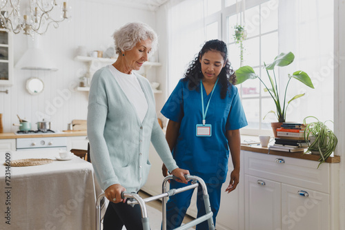 Side view of black african american nurse volunteer teaching elderly woman with gray hair in cardigan to use walker, supporting her and giving advice and instructions, standing in her kitchen photo