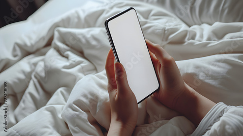 Hand holding an isolated smartphone device with blank empty white screen in bed in the bedroom, business communication technology concept