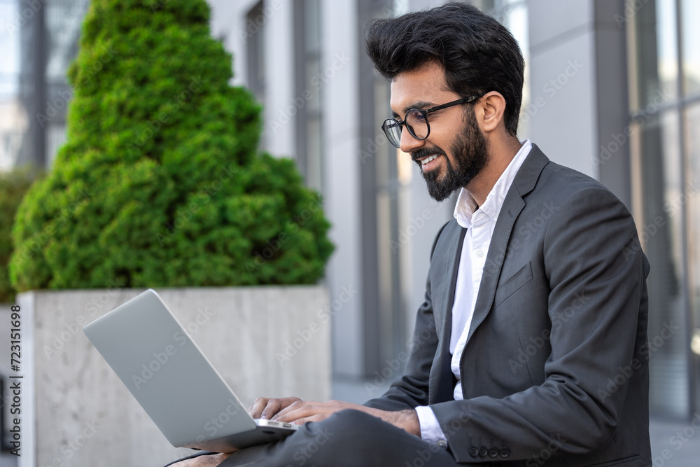 Young hindu businessman working on a laptop while sitting near the office building Young hindu businessman working on a laptop while sitting near the office building