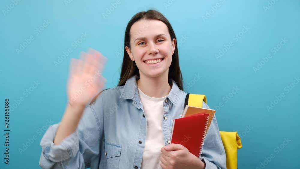 Portrait of charming young woman student wearing shirt with backpack and holding books waving saying hello happy and smiling, friendly welcome gesture, isolated on blue background wall in studio Portrait of charming young woman student wearing shirt with backpack and holding books waving saying hello happy and smiling, friendly welcome gesture, isolated on blue background wall in studio