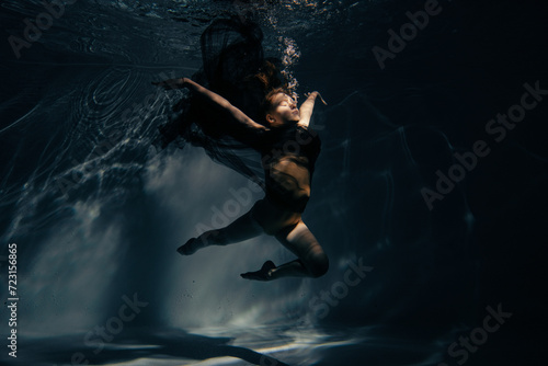 Underwater shoot of ballerina in black dress swimming and dancing in water through sunbeams.