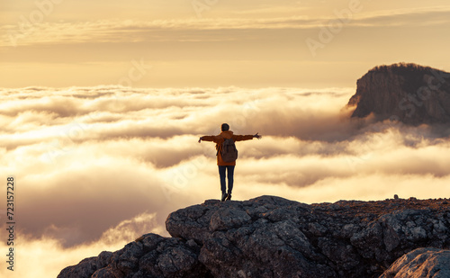 Silhouette of happy woman hiker with backpack is standing with open arms above clouds and enjoys sunset