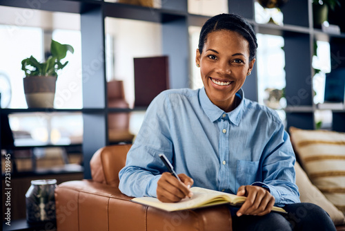 A portrait of a smiling black woman, sitting on the sofa in her office, writing some notes in her notebook. photo