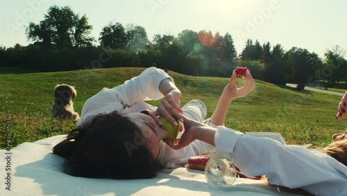 Woman offers friend holding strawberry to eat apple. LGBT couple creates memories spending time in meadow that linger in hearts