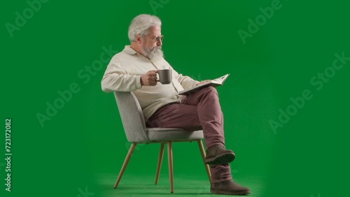 Portrait of aged bearded man on chroma key green screen background. Full shot of senior man sitting on a chair drinking tea reading book.