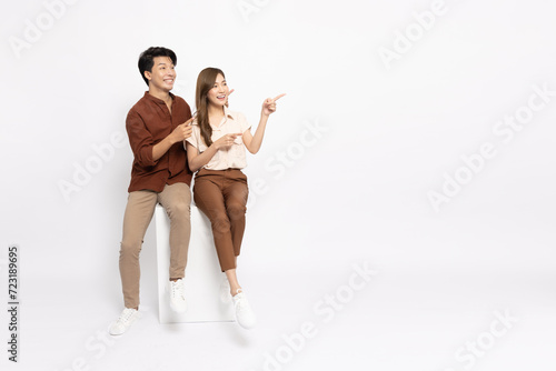 Young Asian couple sitting on white box and pointing to empty copy space isolated on white background, Two people concept