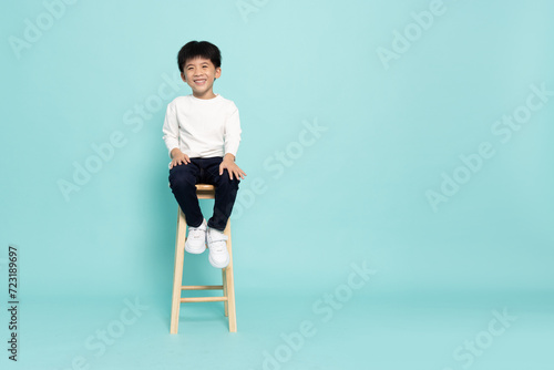 Smiling Asian little boy sitting on wood chair studio isolated on green background, Full body composition