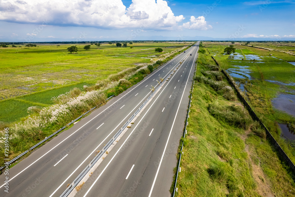 Fototapeta premium Scenic aerial shot of an empty highway cutting through lush green fields under a clear blue sky, emanating tranquility and ease. At Central Luzon Link Expressway.