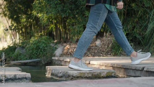 Female legs in jeans and sneakers stepping on stones across a small stream. Slow motion. photo