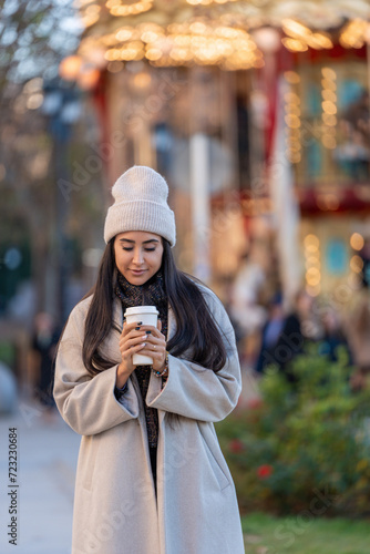 Young lady relishing her coffee on a cold evening, with carousel lights softly glowing in the background. 