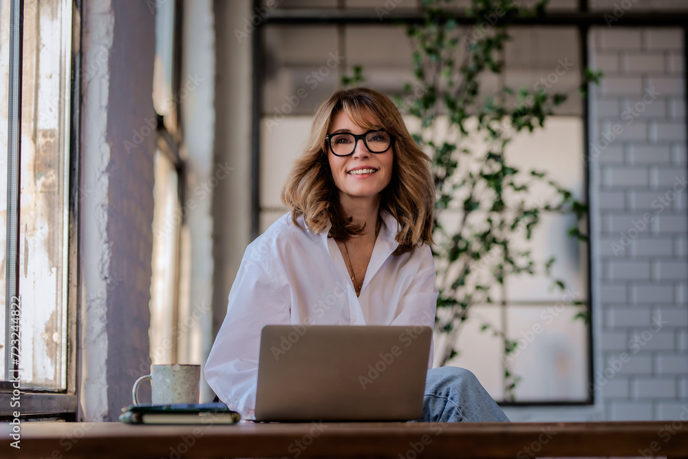 Attractive mid aged woman sitting at the kitchen at home and using her laptop for work Attractive mid aged woman sitting at the kitchen at home and using her laptop for work