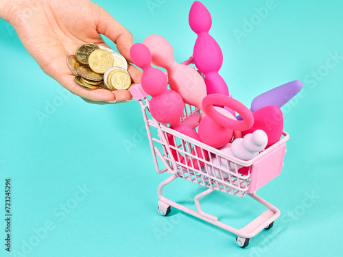 Female hand with heap of euro cents with pink shopping trolley with sex toys in it over turquoise background