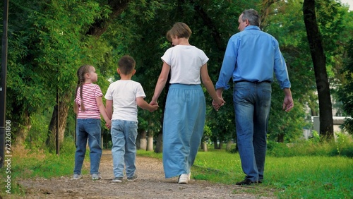 Happy kids stroll clasping hands with lovely parents in park on vacation. Elated parents relish meandering with interconnected hands near kids. Boy and girl amble hand in hand accompanied by parents
