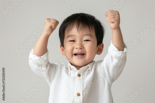 Exultant One-Year-Old Asian Boy In A White Shirt Raises His Fist In Triumph: Perfectly Symmetrical Photo With Centered Composition And Ample Copy Space