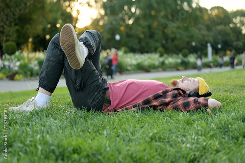 A red-haired guy in a yellow hat lies down on the grass and enjoys a summer day, listening to music on headphones.