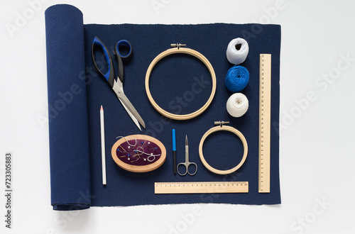 Set of accessories for hand embroidery in Sashiko style: blue canvas, white threads, pincushion with needles, scissors, wooden hoops and rulers on white table. DIY concept. Flat lay, closeup, top view