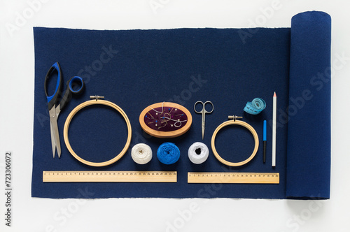 Top view of accessories for hand embroidery in Sashiko style: blue canvas, white threads, pincushion, needles, scissors, wooden hoops, tailor's pencil and rulers on white table. Flat lay, copy space