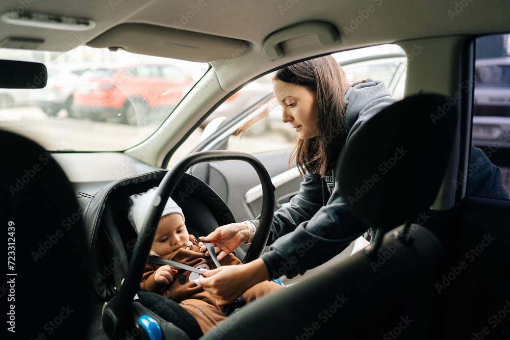 View from backseat of pretty loving mother fasting infant baby boy on front passenger seat with back to engine. Cute toddler with security belt in vehicle. Concept of safety lifestyle, driving, travel
