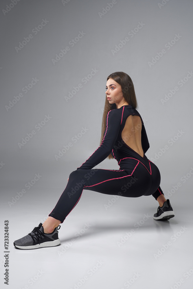 Fitness beauty in black bodysuit doing squatting, while training in studio. View from shoulder of slim dark haired female exercising alone, looking away, isolated on grey background. Concept of sport. Fitness beauty in black bodysuit doing squatting, while training in studio. View from shoulder of slim dark haired female exercising alone, looking away, isolated on grey background. Concept of sport.