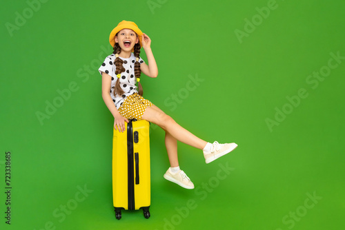 A young girl with a yellow suitcase is going on a trip. A happy teenage girl in a summer hat and shorts is sitting on her luggage and smiling broadly. Green isolated background.