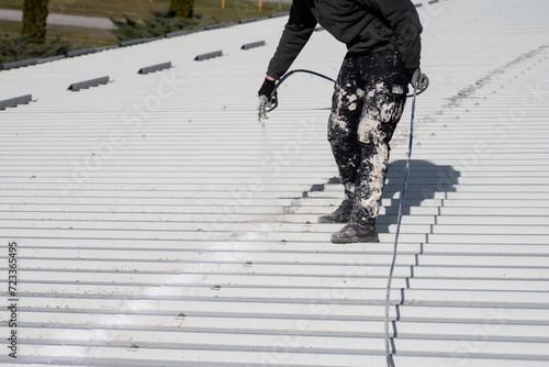 Metal roof painting with compressor. Technician performing painting of roof in white color with high pressure system. Work at height. Roof repairing service. Rooftop maintenance.