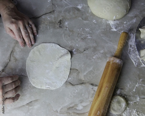on the kitchen table, women's hands roll out dough with a rolling pin. several round pieces of dough and a prosky piece for stuffing it with meat are visible.