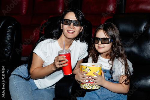Cute cheerful little beautiful girl eating popcorn enjoying watching a cartoon at the cinema with her mother, family happiness movies entertainment parenting childhood emotions. High quality photo.