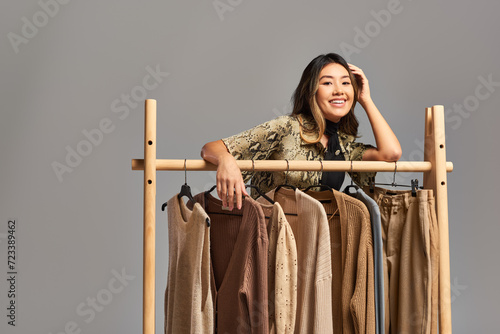 cheerful asian stylist smiling at camera near rack with bespoke clothes on grey, fashion business
