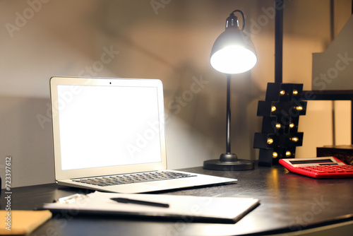 Blank laptop with clipboard and glowing lamp on table in office at night