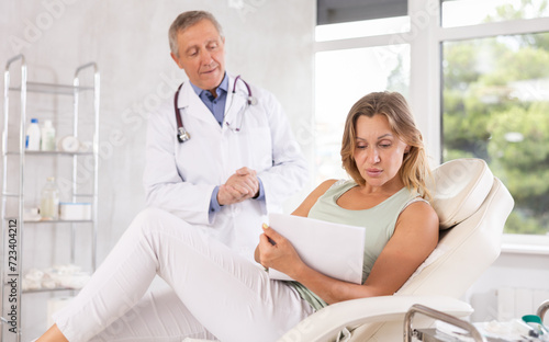Middle-aged female patient lying on medical chair signing papers while talking with man doctor