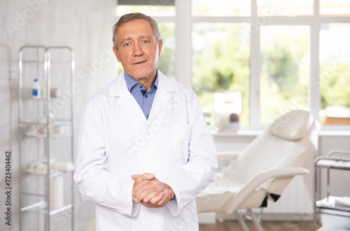 Positive old man doctor posing against background of doctor s cabinet with clinical chair