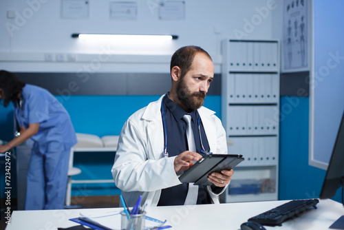 Male doctor reading patient records while holding a tablet. In the clinic office, caucasian man wearing lab coat is using digital gadget and desktop pc to review medical information.