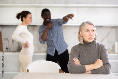 Angry mature woman sitting at table while two male and female family members disputing with each other in the back