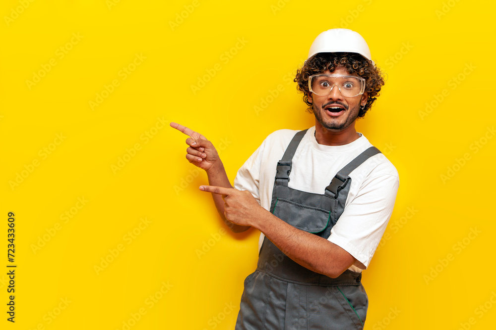 Young shocked Indian builder in hard hat and overalls points with hands to the side on yellow isolated background, surprised Indian foreman in uniform showing and advertising copy space in amazement - obrazy, fototapety, plakaty 