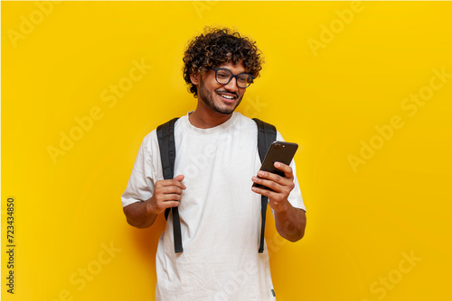 young indian guy student in a white t-shirt with glasses and with a backpack uses a smartphone and selects online on a yellow isolated background, a curly-haired man types a message on a mobile