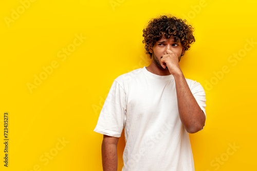 young curly indian man covering his nose with his hand and showing disgust on a yellow isolated background, displeased indian man sniffing an unpleasant smell photo