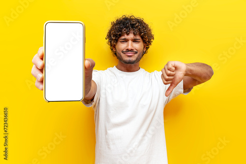sad dissatisfied Indian man showing blank smartphone screen and dislike on yellow isolated background, curly guy advertising phone online and showing bad review photo