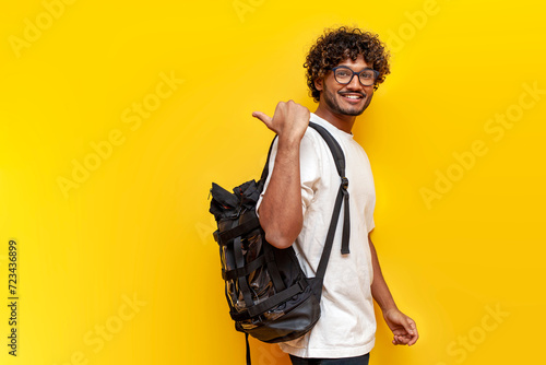 young indian guy student in a white t-shirt with glasses and with backpack points back on yellow isolated background, curly man goes to college to study and points at the copy space from behind photo